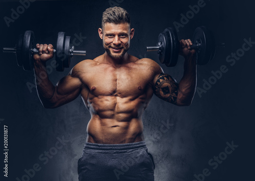 A handsome shirtless tattooed bodybuilder with stylish haircut and beard, wearing sports shorts, posing in a studio. Isolated on a dark background © Fxquadro