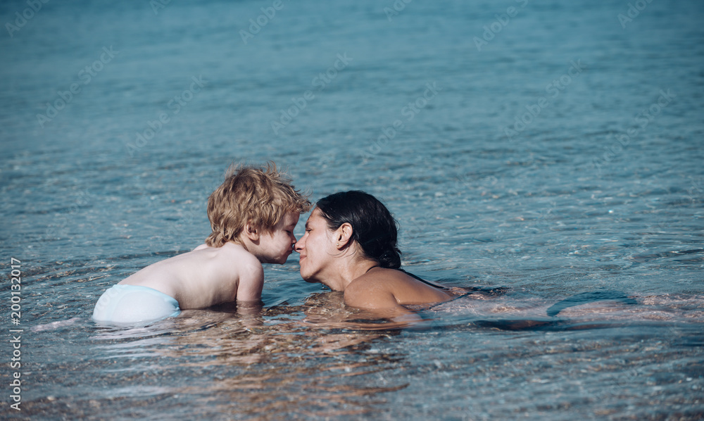 Mom and kid with smiling faces spend time together in sea on sunny day. Cute child boy play with mom in sea water, touch her nose. Motherhood concept. Mother swimming in sea and look at child. Mom and kid with smiling faces spend time together in sea on sunny day. Cute child boy play with mom in sea water, touch her nose. Motherhood concept. Mother swimming in sea and look at child.
