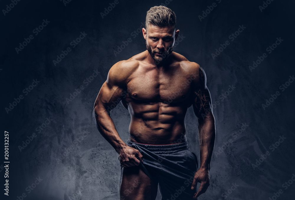 A handsome shirtless tattooed bodybuilder with stylish haircut and beard,  wearing sports shorts, posing in a studio. Isolated on a dark background  Stock Photo | Adobe Stock