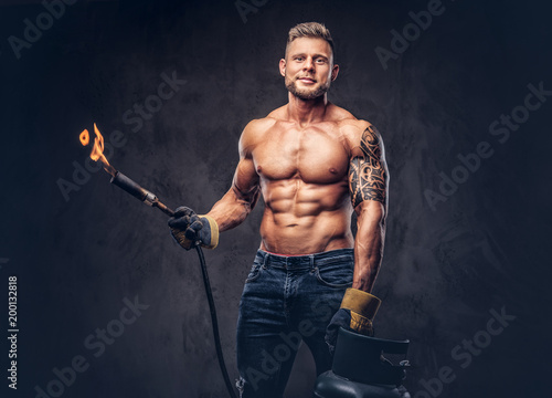 Bild auf Leinwand Brutal tattoed male welder with a stylish haircut and beard, with muscular body, dressed in only jeans, holds propane tank and a burning burner, standing in a studio, looking at a camera