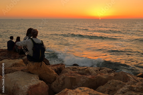 Zakochana para, chłopak i dziewczyna, przytuleni, siedzą na skalistym brzegu morza i podziwiają romantyczny zachód słońca