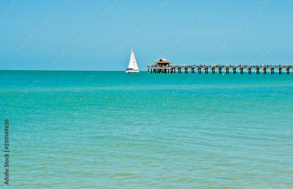 Obraz premium Long view of a sailboat under way, with filled sails passing a wood pier jutting out from a tropical beach with calm waters of the Gulf of Mexico