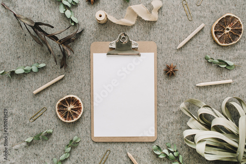 Clipboard with blank paper mock up in frame of eucalyptus branches, dried oranges and leaves, ribbon on beige blanket background. Flat lay, top view wedding rustic concept.