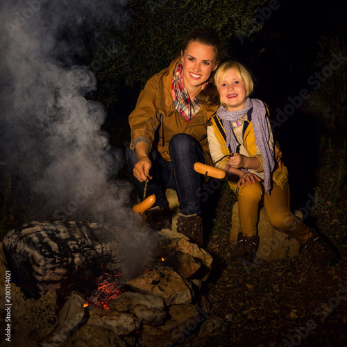 mother and daughter travellers near campfire grilling sausages