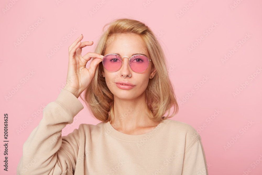 Young blonde woman, shy and smile, stares through spectacles, wears yellow sweatshirt. Isolated over pink background