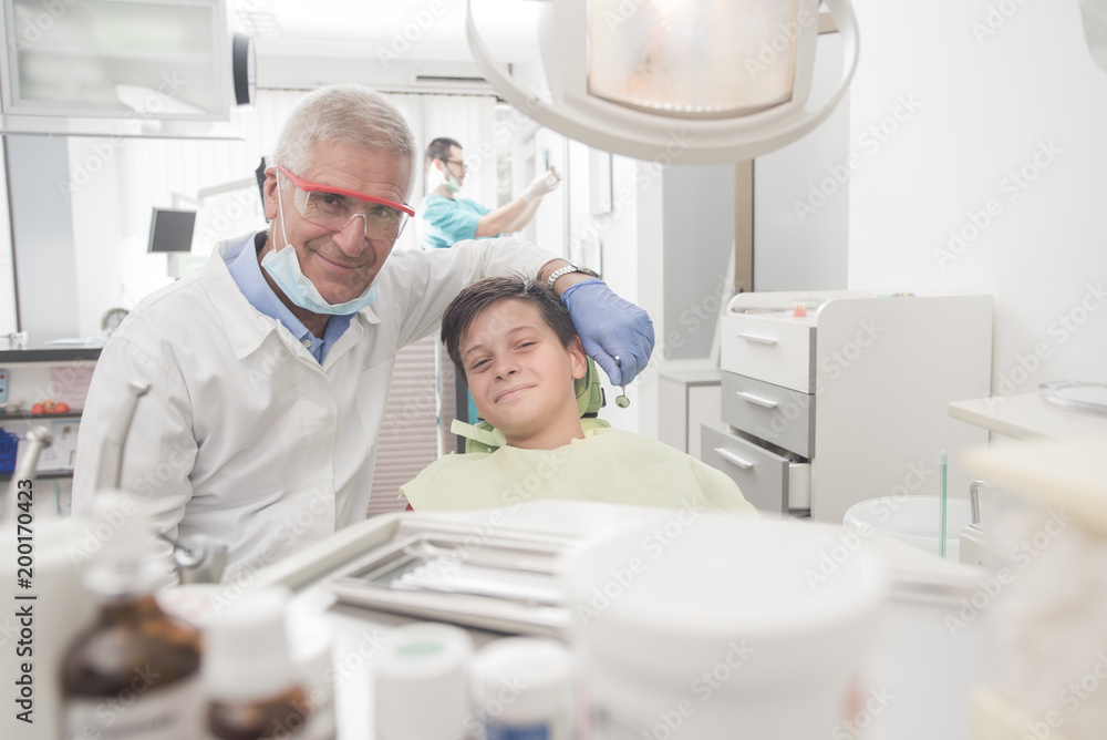 Obraz premium Boy with perfect teeth at the dentist doing check up while other doctors analyze x-ray in the background - oral hygiene health care concept