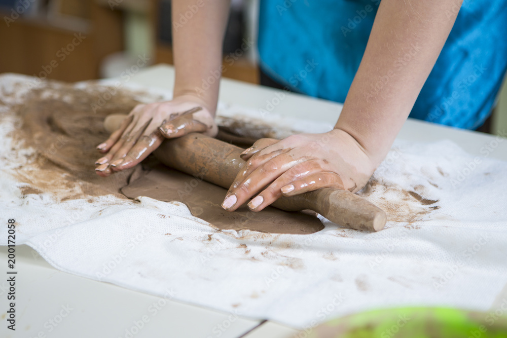 Pottering Ideas and Concepts. Extreme Closeup of Hands of Female Professional in Apron Rolling Out a Piece of Clay with Rolling Pin in Workshop. Pottering Ideas and Concepts. Extreme Closeup of Hands of Female Professional in Apron Rolling Out a Piece of Clay with Rolling Pin in Workshop.