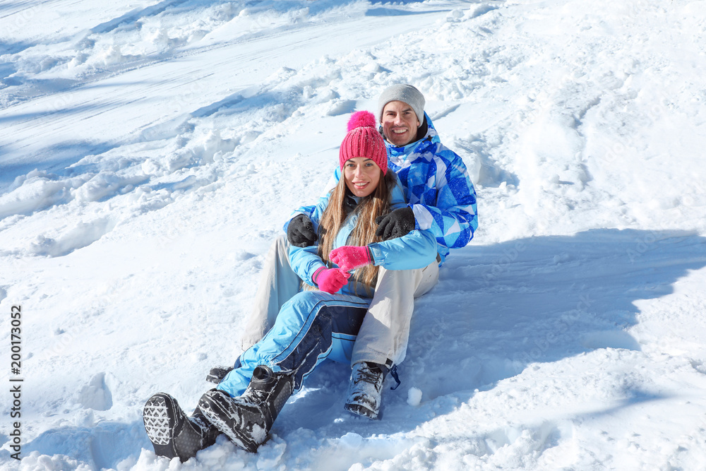 Happy couple sitting on snow at resort on sunny day. Winter vacation