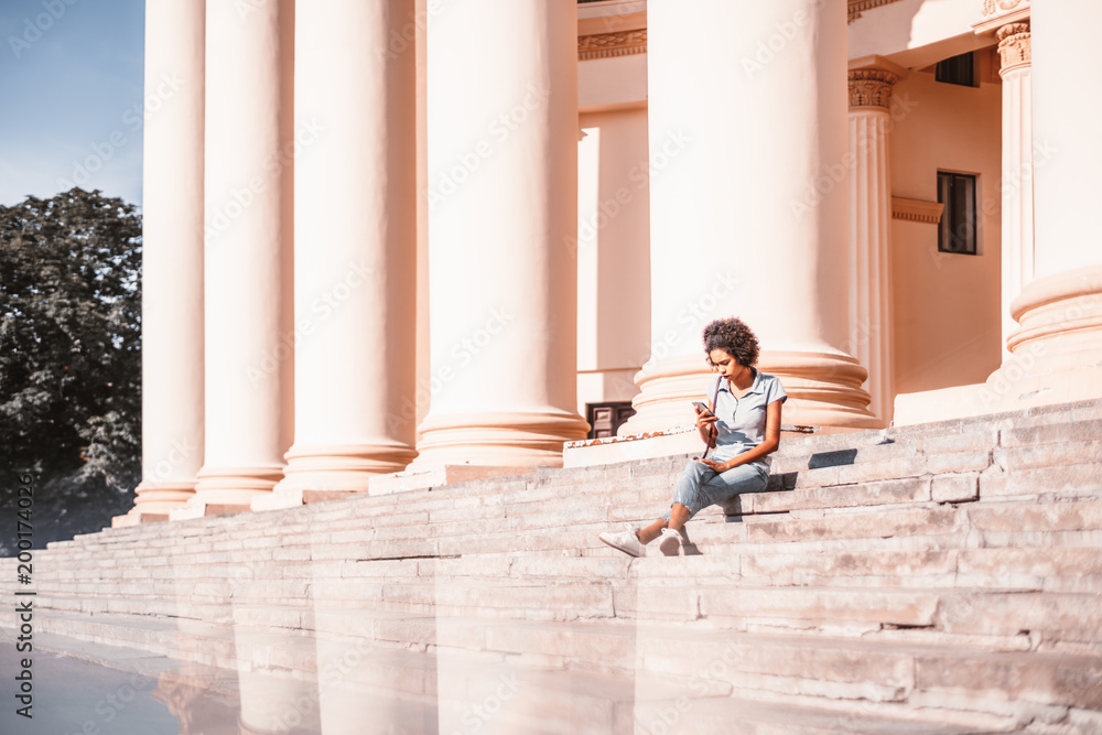 Young curly black female with Afro hair is sitting on the concrete stairs with next to a building with the huge pillars, and texting message to her friend via smartphone; strong reflections below