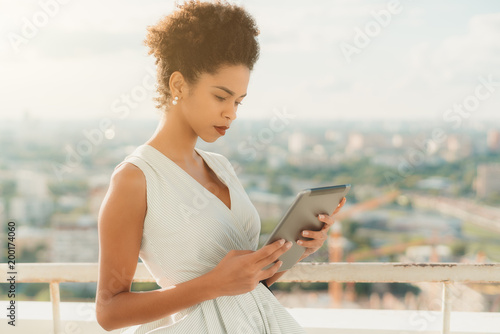 A serious young appealing African-American girl in a white dress is calling her boyfriend via tablet pc to show him a beautiful view of the cityscape from observation area of a skyscraper