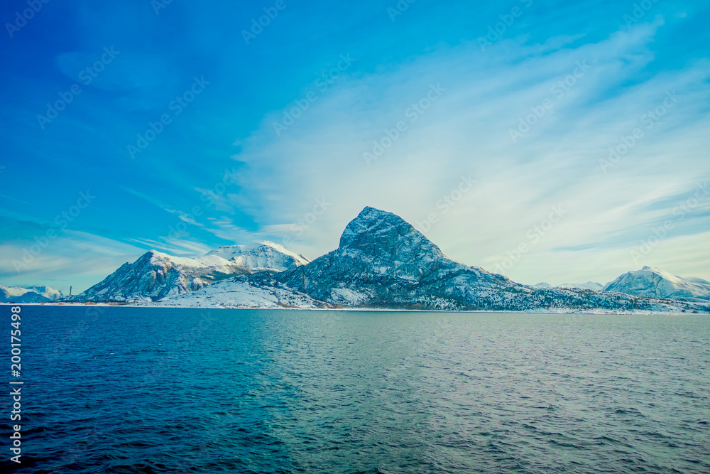 Outdoor view of coastal scenes of huge mountains partial covered with snow durig a trip in Hurtigruten in a blue sky