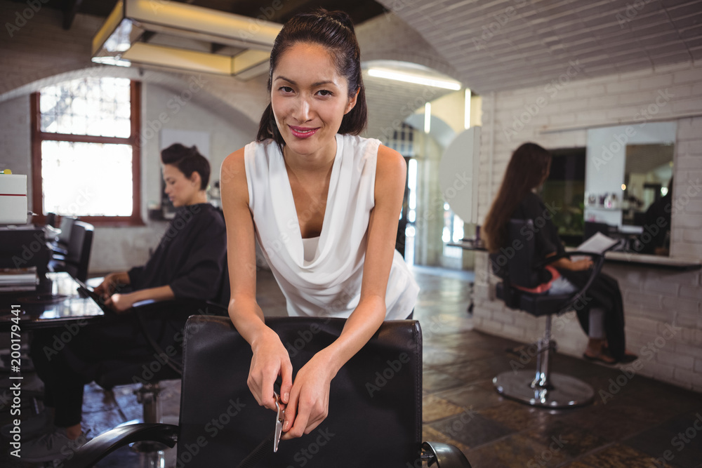Smiling female hair dresser leaning on chair at salon Smiling female hair dresser leaning on chair at salon