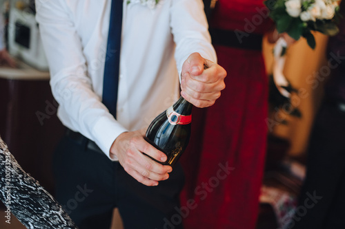 A young man groom in a white shirt opens a bottle of champagne.