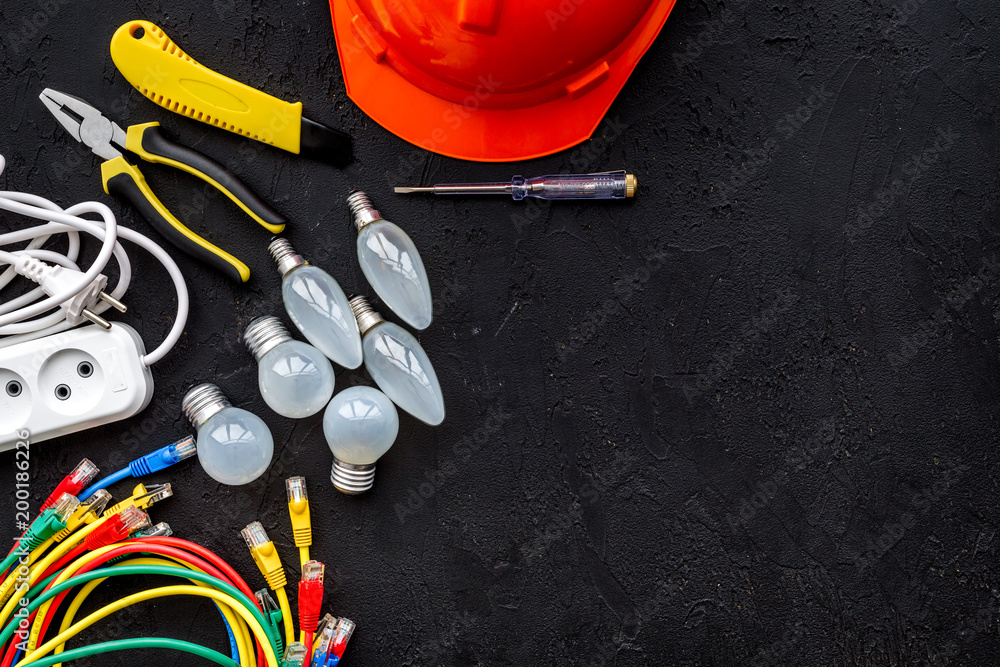 Electrician work concept. Hard hat, tools, cabel, bulb, socket outlet on black background top view space for text