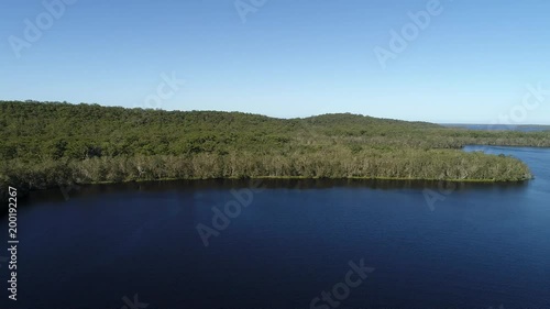 Blue water of Myall lake and feeding streams in aerial flying and rising from above woods level higher in the sky overlooking distant waterfront and pacific ocean.
 photo