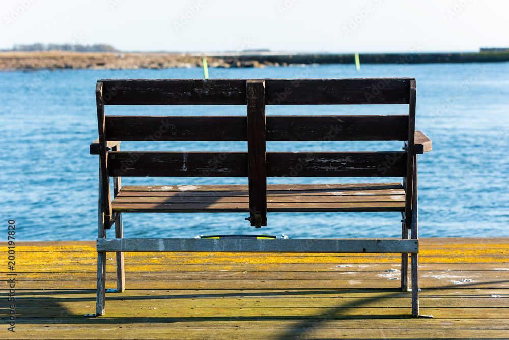 Empty wooden bench at the end of a pier with a blurred view of harbor inlet and coastland. Location Gronhogen on Oland, Sweden.
