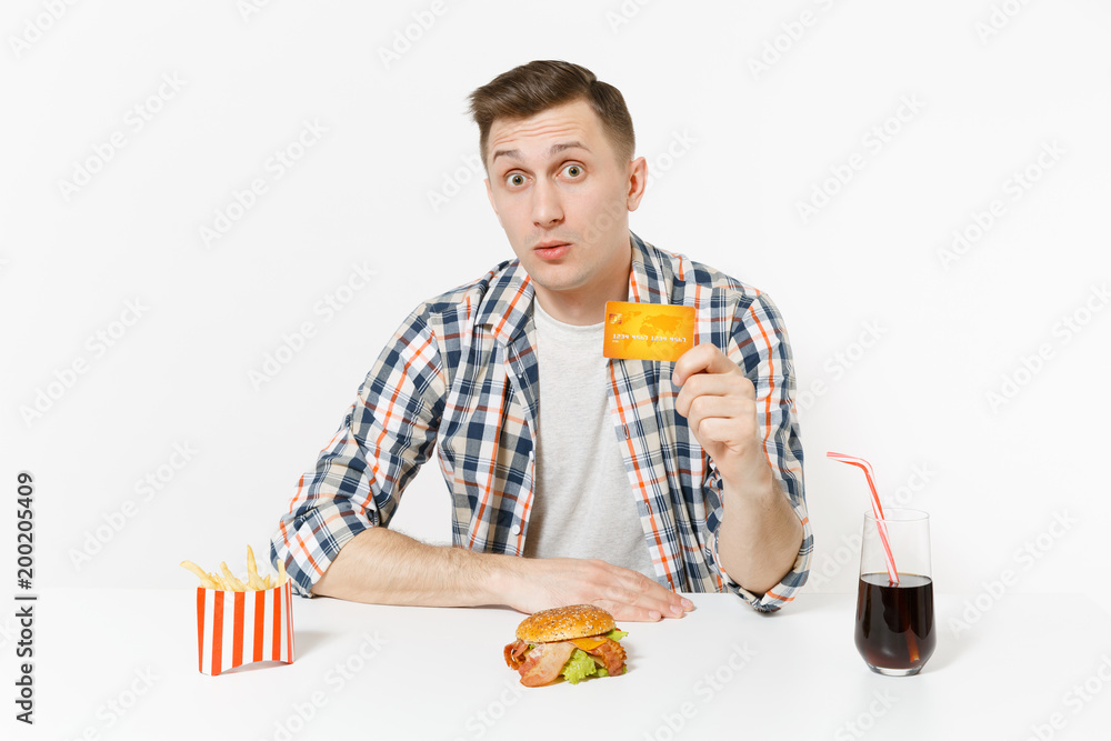 Handsome young man sitting at table with credit card, burger, french fries, cola in glass isolated on white background. Proper nutrition or American classic fast food. Advertising area with copy space Handsome young man sitting at table with credit card, burger, french fries, cola in glass isolated on white background. Proper nutrition or American classic fast food. Advertising area with copy space