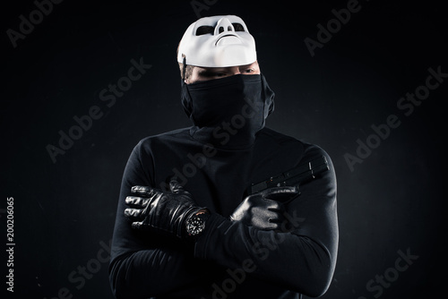 Thief in black balaclava and white mask holding gun in folded arms on black