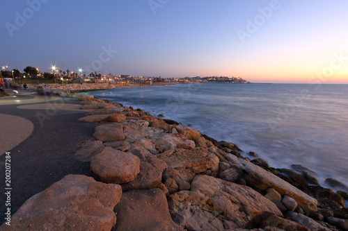 Fototapeta Naklejka Na Ścianę i Meble -  Promenada nadmorka w Tel Awiwie, Izrael, kamienie umacniające brzeg, morze, światła latarń ulicznych, w oddali, na horyzoncie, widoczna Hajfa, wieczór, długie naświetlanie