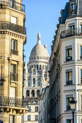 The Basilica of the Sacred Heart in Paris seen through a narrow street between typical buildings with a vintage street light in the foreground under a clear blue sky.
