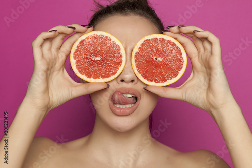 Young sexy woman posing with slices of red grapefruit on her face on pink background