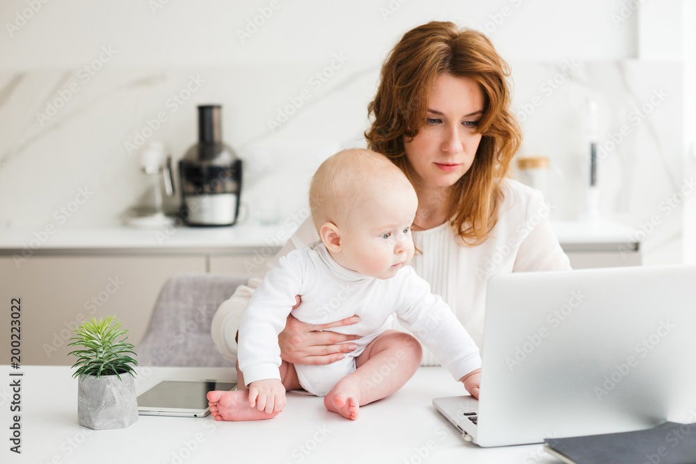 Thoughtful business woman sitting at the table and working on laptop while holding her cute little baby near isolated Thoughtful business woman sitting at the table and working on laptop while holding her cute little baby near isolated