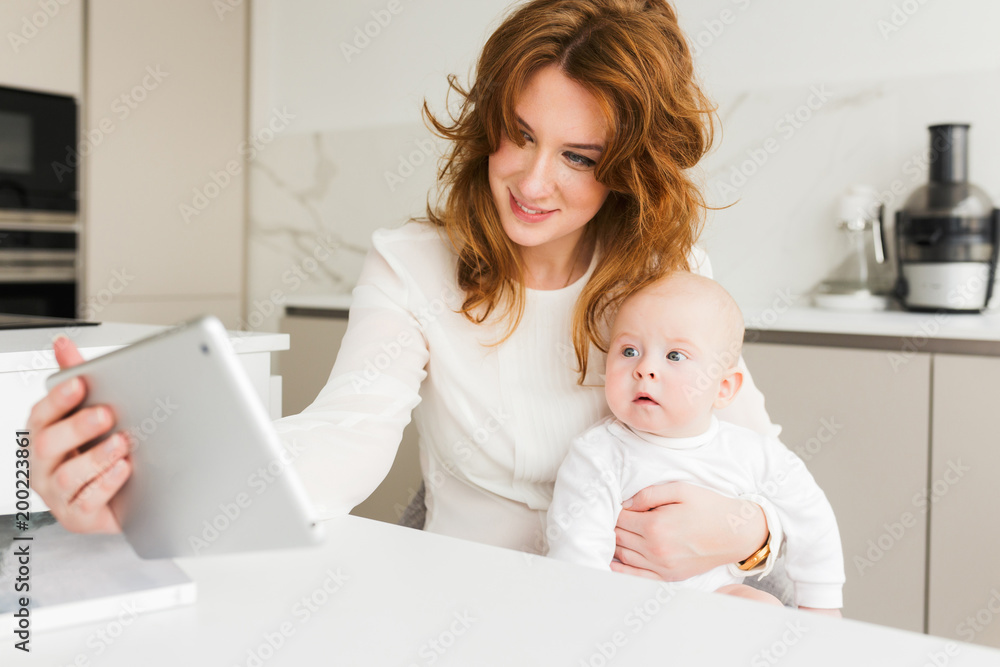 Portrait of smiling woman sitting and holding her cute little baby in hand while happily using her tablet isolated Portrait of smiling woman sitting and holding her cute little baby in hand while happily using her tablet isolated