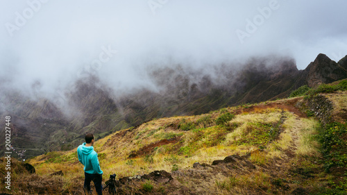 Fototapeta Naklejka Na Ścianę i Meble -  Panoramic shot of traveler taking picture of amazing steep mountain range and terrain canyon valley on the path from Xo-Xo Valley. Camera on tripod.Santo Antao Island, Cape Verde