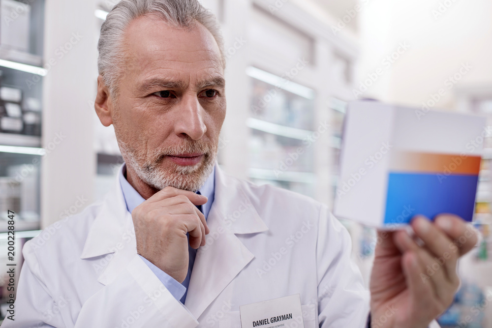 Right dosage. Senior attentive male pharmacist touching chin while rising medication Right dosage. Senior attentive male pharmacist touching chin while rising medication