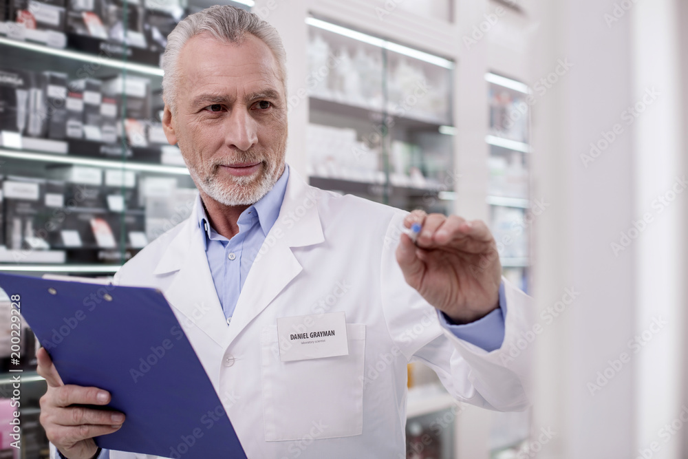 Medication list. Senior handsome male pharmacist stretching hand while using clipboard