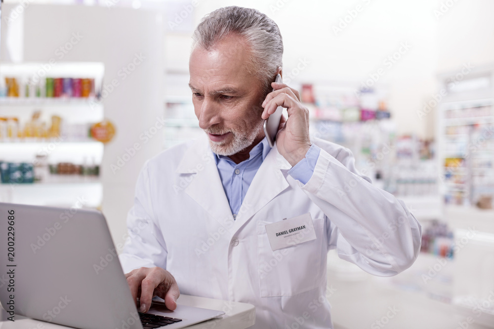 Business call. Senior attentive male pharmacist working on laptop while calling Business call. Senior attentive male pharmacist working on laptop while calling