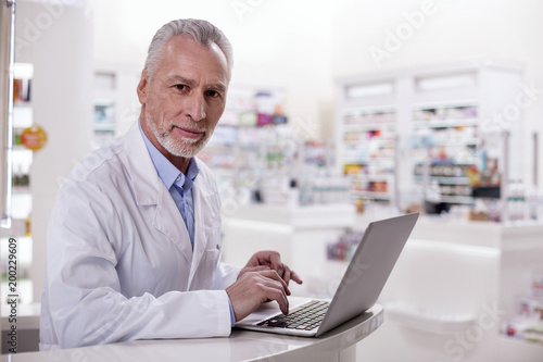 Pharmacy organization. Mature earnest male pharmacist working on laptop while looking at camera