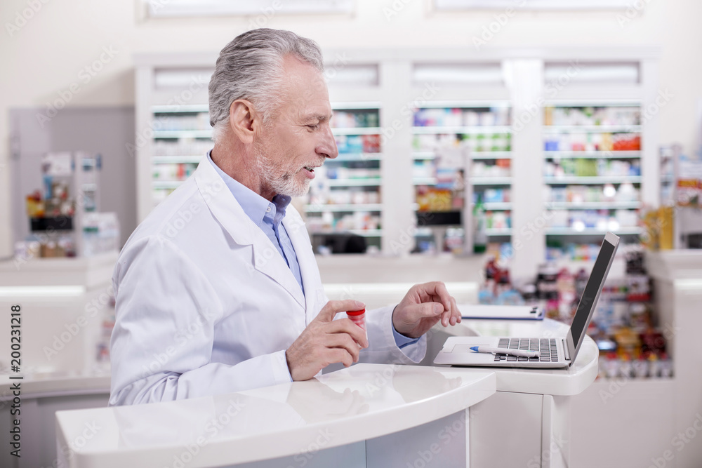 Let me check. Senior attentive male pharmacist posing in profile while typing on laptop Let me check. Senior attentive male pharmacist posing in profile while typing on laptop