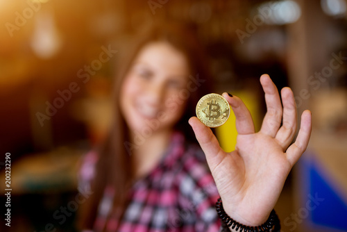 Young cheerful adorable girl is extending her arm toward the camera and showing a bitcoin.