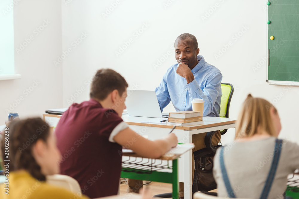 Writing schoolchildren and teacher sitting at table with laptop and coffee cup