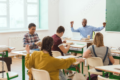 Multicultural group of teenage schoolchildren and smiling african american teacher with raising arms