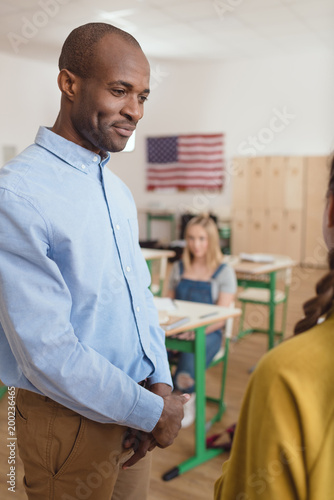 Smiling african american teacher talking to schoolgirl and classmate sitting behind in classroom with flag of USA