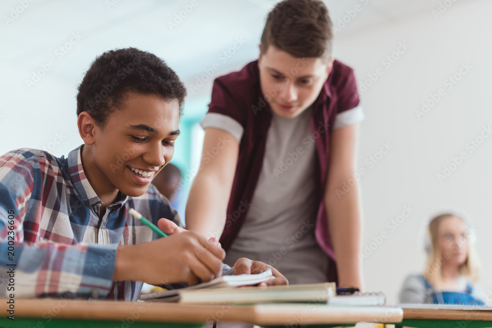 Smiling african american boy writing in textbook with classmate standing behind Smiling african american boy writing in textbook with classmate standing behind