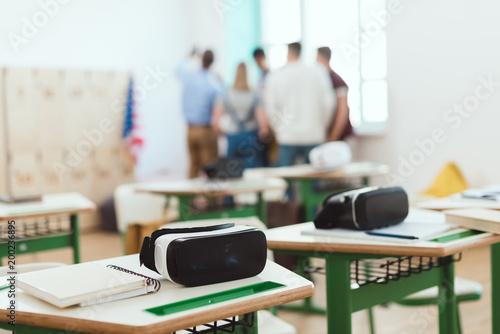 Virtual reality headsets on tables with teacher and high school students standing behind in classroom
