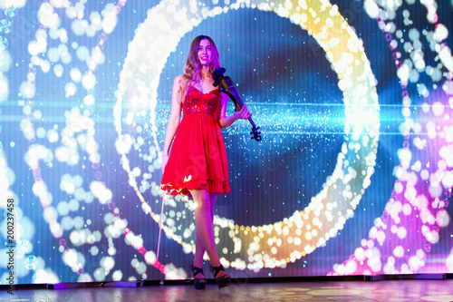 young sexy girl violoncellist in red dress is standing fashion and playing the cello on the blue light screen background