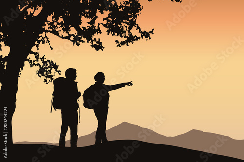Two tourists standing under a tree pointing in the distance in mountain landscape with orange sky and space for your text