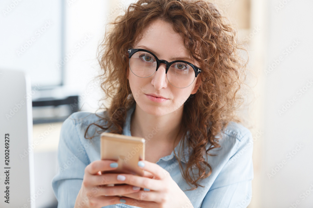 Concentrated serious female in round eyewear holds smart phone in hands and communicates remotely with partners, creates publication work Concentrated serious female in round eyewear holds smart phone in hands and communicates remotely with partners, creates publication work