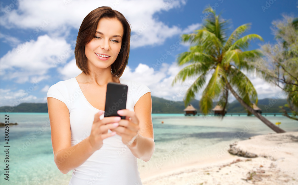 travel, tourism, summer holidays and vacation concept - happy young woman taking selfie by smartphone over exotic tropical beach with palm tree and bungalow sheds background