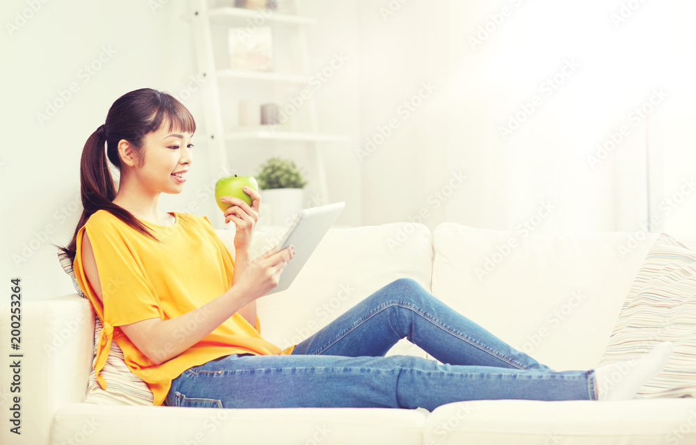 people, healthy eating, education, technology and concept - happy young asian woman sitting on sofa with tablet pc computer and green apple at home