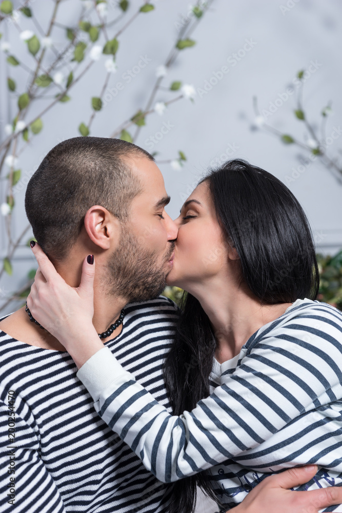 Charming international couple in striped sweaters kissing and hugging while sitting on the bed Charming international couple in striped sweaters kissing and hugging while sitting on the bed