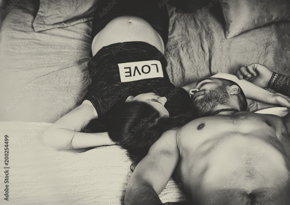 Black and white photo of wonderful international couple of man with bare chest and brunette pregnant woman look at each other while lying on the bed
