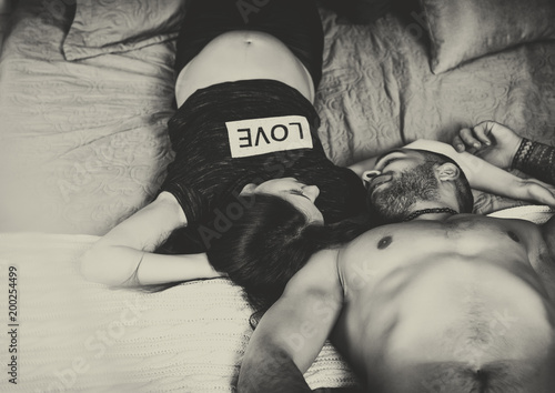 Obraz na płótnie Black and white photo of wonderful international couple of man with bare chest and brunette pregnant woman look at each other while lying on the bed w salonie