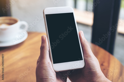 Mockup image of hands holding white mobile phone with blank black screen and coffee cup on wooden table in cafe