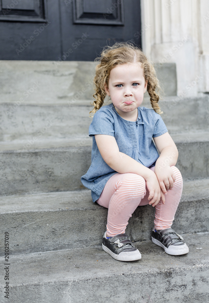 Emotional portrait of a sad, unhappy little girl with blond curly hair sitting on the front steps of her home in an urban setting. Cute expression and adorable face. child behavior concept photo Emotional portrait of a sad, unhappy little girl with blond curly hair sitting on the front steps of her home in an urban setting. Cute expression and adorable face. child behavior concept photo