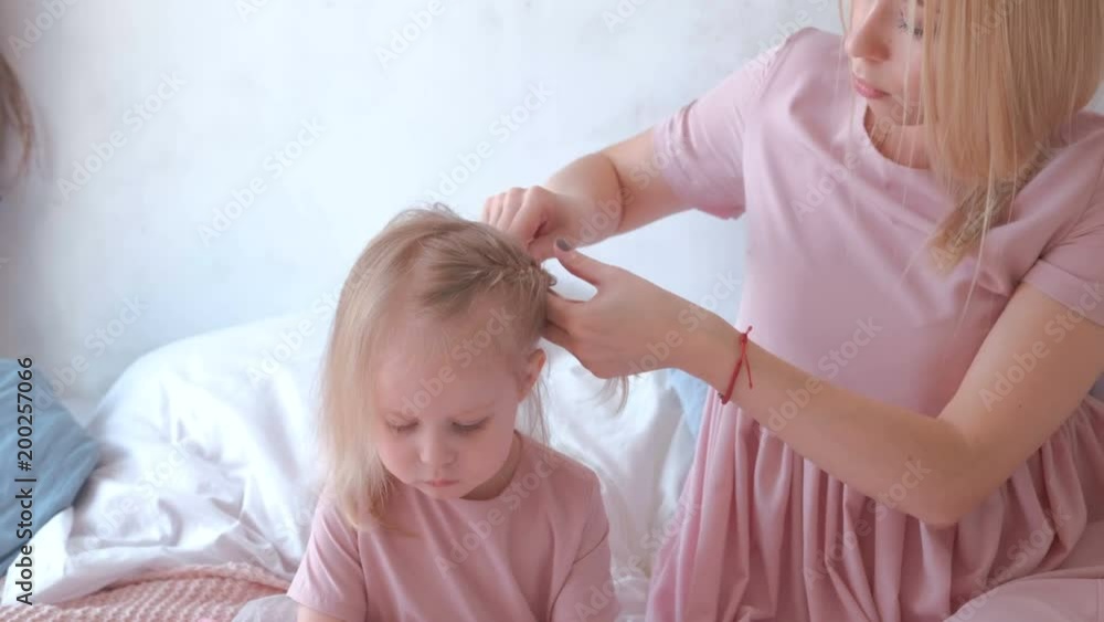 Young attractive blond mom tie hair into braids to her little charming daughter in pink dresses while she painting with felt-pen Young attractive blond mom tie hair into braids to her little charming daughter in pink dresses while she painting with felt-pen
